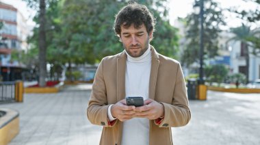 Sıradan iş kıyafetleri içinde yakışıklı bir İspanyol, şehir caddesinde yeşilliklerle çevrili dururken akıllı telefonuyla mesajlaşmaya dalmış..