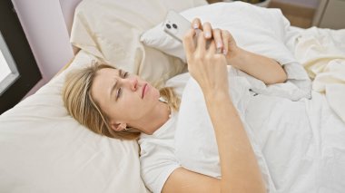 Genç sarışın bir kadın yatak odasında akıllı telefonunu karıştırarak yatakta uzanıyor..