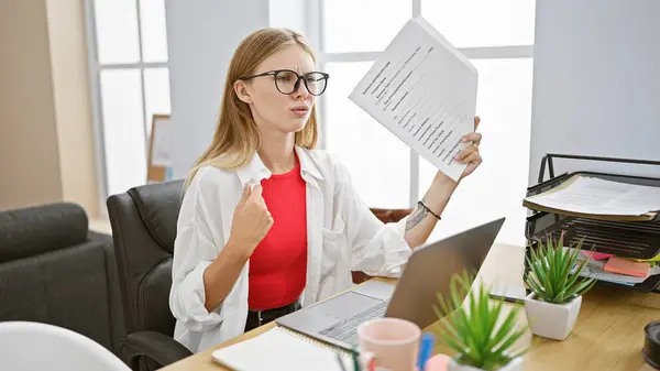 stock image Blonde woman in office fanning herself with paper, looking tired, amidst work documents and laptop.