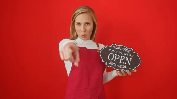 stock image A young caucasian woman in a red apron points at the camera while holding an 'open' sign against a solid red background.