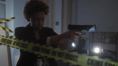 Silahlı Afrikalı Amerikalı kadın dedektifler bantla işaretlenmiş loş ışıkta suç mahallini inceliyor.