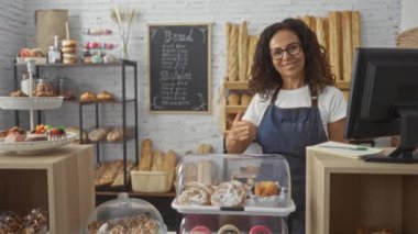 Fırında çalışan kadın, arka planda ekmeği olan taze hamur işlerini gösteriyor. Gülümsüyor ve tezgahın arkasında duruyor.