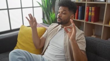 Yakışıklı Afro-Amerikan adam yoga pozisyonunda meditasyon yapıyor. Kanepede, gözleri kapalı, nefes alıyor.