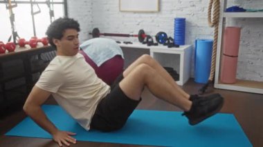 Spor salonundaki bir paspasın üzerinde, spor aletleri ve ağırlıklarla dolu bir yerde karın egzersizi yapan genç adam..