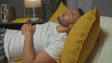 Beyaz tişörtlü, sakallı genç bir İspanyol rahat bir yatak odasında sarı bir yastıkta dinleniyor..