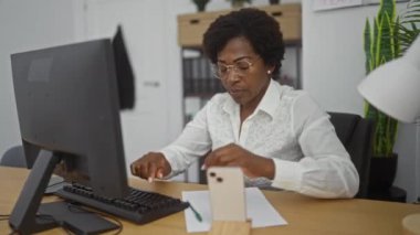 Bilgisayarda çalışan bir kadın, arka planda bitki ve raflar olan ofis çalışma alanındaki bir masada akıllı telefonuyla çalışıyor.