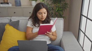 Genç bir İspanyol kadın oturma odasında oturmuş kitap okuyor ve dizüstü bilgisayar kullanıyor. Modern, rahat bir ev ortamını somutlaştırıyor..