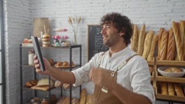 Genç bir adam fırında video görüşmesi yapıyor, ev fırını, tablet kullanıyor, arka planda ekmek ve pastalarla dolu, İspanyol asıllı, erkek fırıncı