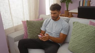 Yakışıklı İspanyol bir adam oturma odasında rahat bir koltukta oturmuş zafer işareti yaparken selfie çekiyor. Etrafı rahat yastıklar ve modern dekorlarla çevrili..