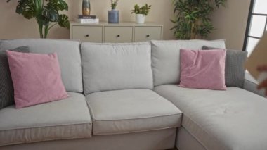 Çiçekler, pembe yastıklar ve tarafsız dekorlarla dolu rahat bir oturma odasında gri bir koltukta oturan İspanyol genç adam bir paket tutuyor..