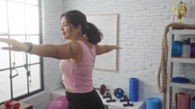 Pembe bluzlu, genç, güzel bir İspanyol kadın, kollarını açık bir spor salonunda geriyor, spor aletleri ve beyaz tahtalarla çevrili..