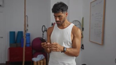 Genç adam arka planda fitness ekipmanıyla spor salonunda telefon ve kasetleri kontrol ediyor.