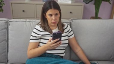 Genç bir İspanyol kadın, oturma odasındaki koltukta elinde telefonla oturuyor. Üzerinde çizgili bir gömlek ve mavi bir battaniye var..