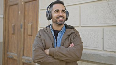 Sakallı yakışıklı, şehir ortamında kulaklıkla müzik dinleyen, duvara yaslanan bir İspanyol.