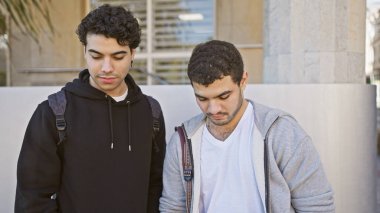 Gündelik giyinmiş iki İspanyol adam şehir sokağında dikiliyorlar, akıllı bir telefona takılıp kalmışlar..