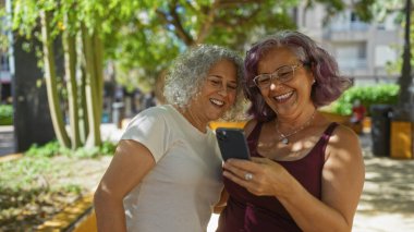 Kıvırcık saçlı iki yaşlı kadın güneşli bir günde açık hava ortamında akıllı telefona bakmaktan hoşlanırlar..