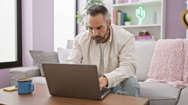 Sakallı, gri saçlı yaşlı bir İspanyol rahat bir oturma odasında dizüstü bilgisayarla çalışıyor..