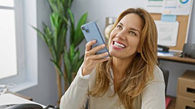 Modern ofiste akıllı telefon kullanan gülümseyen bir kadın kapalı bir şirket ortamında bağlantı ve profesyonelliği resmediyor..