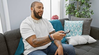 Evde elinde uzaktan kumandayla koltukta oturan sakallı rahat bir adam..