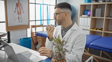 Beyaz laboratuvar önlüklü düşünceli bir İspanyol, fizyoterapi kliniğindeki laptopta hasta verilerini değerlendiriyor..