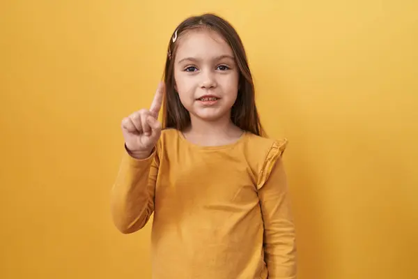 stock image Adorable hispanic girl, confidently pointing one finger up with a happy face, standing isolated on a yellow background, ready to share a smart idea or thoughtful question.