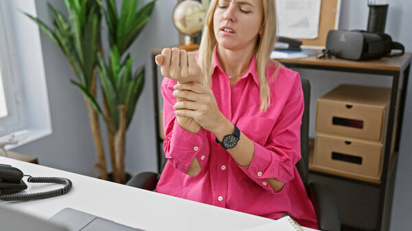 A woman in a pink shirt experiences wrist pain in an office setting, indicating potential work-related strain.