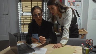 İki kadın, iyi organize edilmiş bir ofis ortamında akıllı telefon ve dizüstü bilgisayar kullanarak bir davayı araştırıyor..