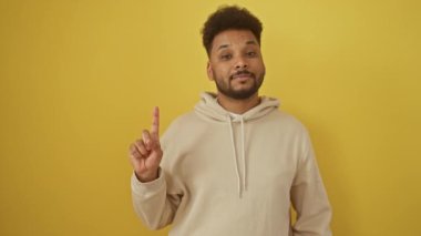 Mutlu siyah adam, ışıl ışıl gülümsüyor ve güvenle bir parmağını yukarı kaldırıyor, sarı kapüşonlu giyiyor. Sarı arka planda izole edilmiş, pozitifliği kişileştiren ve bir numara olan.