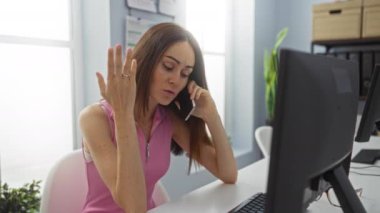 Modern bir ofis ortamında, bilgisayarın yanında kolsuz gömlek giyen bir kadın telefonda stresli görünüyor.
