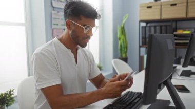 Modern mobilya ve dekorasyonlarla çevrili bilgisayar masasında akıllı telefon kullanan genç bir adam.