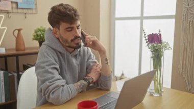 Sakallı genç bir İspanyol, rahat bir dairede dizüstü bilgisayarla çalışıyor. Telefonuyla konuşuyor..