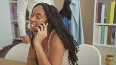 Bir kadın telefonda konuşuyor, rahat bir oturma odasında kendisine sarılan bir adama gülümsüyor, evde sevgi ve iletişimi vurguluyor..