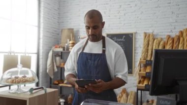 Bir fırında çalışan genç adam önlük takıyor ve etrafında ekmek rafları ve pastalarla dolu bir tablet kullanıyor..