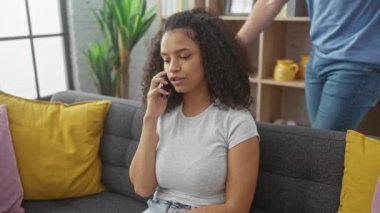 Bir erkek, kanepede gülümseyen bir kadına şefkatle sarılır. O da rahat bir oturma odasında telefonla konuşur..