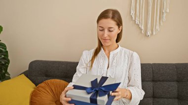 Evdeki kanepede oturan gülümseyen genç bir kadın elinde büyük mavi bir kurdeleyle nazikçe bir hediye tutuyor..