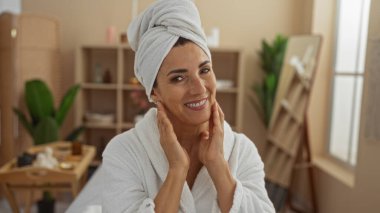 Kaplıcada rahatlatıcı bir anın tadını çıkaran bir kadın, beyaz bir bornoz giymiş, başının etrafına bir havlu sarılmış, dingin ve iyi dekore edilmiş bir sağlık merkezi ile çevrili..