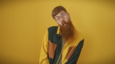 Sakallı, gözlüklü ve renkli gömlekli genç adam sarı arka planda poz veriyor..