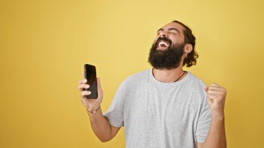Sakallı, mutlu İspanyol adam sarı arka planda akıllı telefonla başarıyı kutluyor.