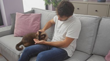 İspanyol bir adam, rahat bir oturma odasında, gri bir kanepede bir siyam kedisini nazikçe okşuyor..