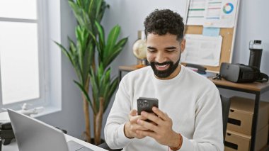 Modern ofis ortamında akıllı telefon kullanan gülümseyen sakallı adam.
