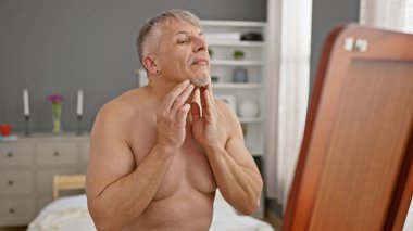 Tıraş köpüğü süren, iyi aydınlatılmış bir yatak odasındaki aynaya bakan, olgun, üstsüz bir adam..