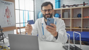 Bir klinikte elinde kredi kartı ve akıllı telefon olan yetişkin bir doktor çevrimiçi ödeme veya teletıp gösteriyor..