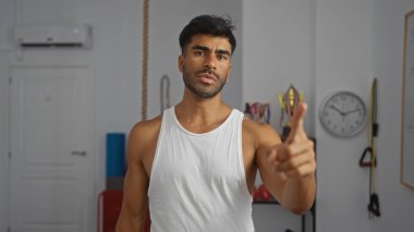 Spor salonunda beyaz atlet giyen İspanyol bir adam önde, içeride spor aletleri ve arkasında bir saatle dikiliyor..