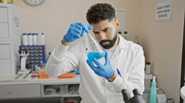 Yakışıklı sakallı adam tıbbi laboratuvar ortamında mavi bir sıvıyla deneyler yapıyor. Eldiven ve laboratuvar önlüğü giyiyor..