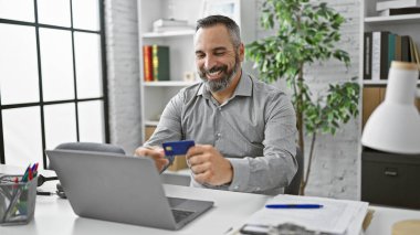 Gri sakallı, gülümseyen adam modern ofiste kredi kartı ve laptop kullanıyor.