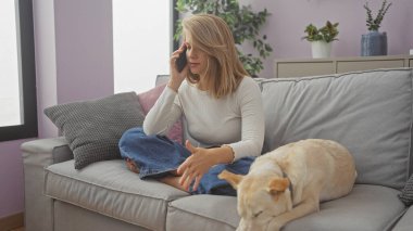 Genç bir kadın, rahat bir oturma odasında dinlenen köpeğinin yanında bir kanepede otururken telefonla konuşur..