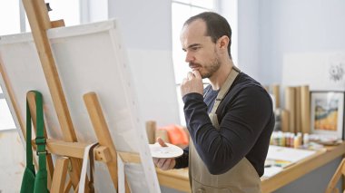 Sanat atölyesinde sakallı, dalgın ve kel bir adam elinde boya paletiyle tuvalin üzerinde kafa yoruyor..