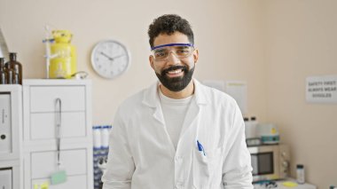 Laboratuvarda gülümseyen laboratuvar önlüklü, sakallı yakışıklı bir genç.