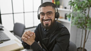Neşeli, sakallı bir adam bilgisayar masasında kulaklık takıyor. Parlak, modern bir ofiste, güvenle gülümsüyor..