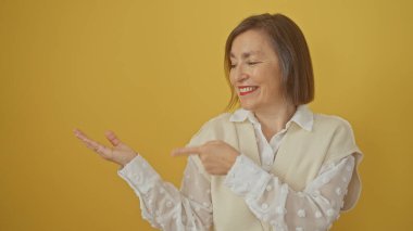Gülümseyen orta yaşlı bir kadın, elini sarı arka plana uzatarak pozitiflik ve kendine güveni yansıtıyor..